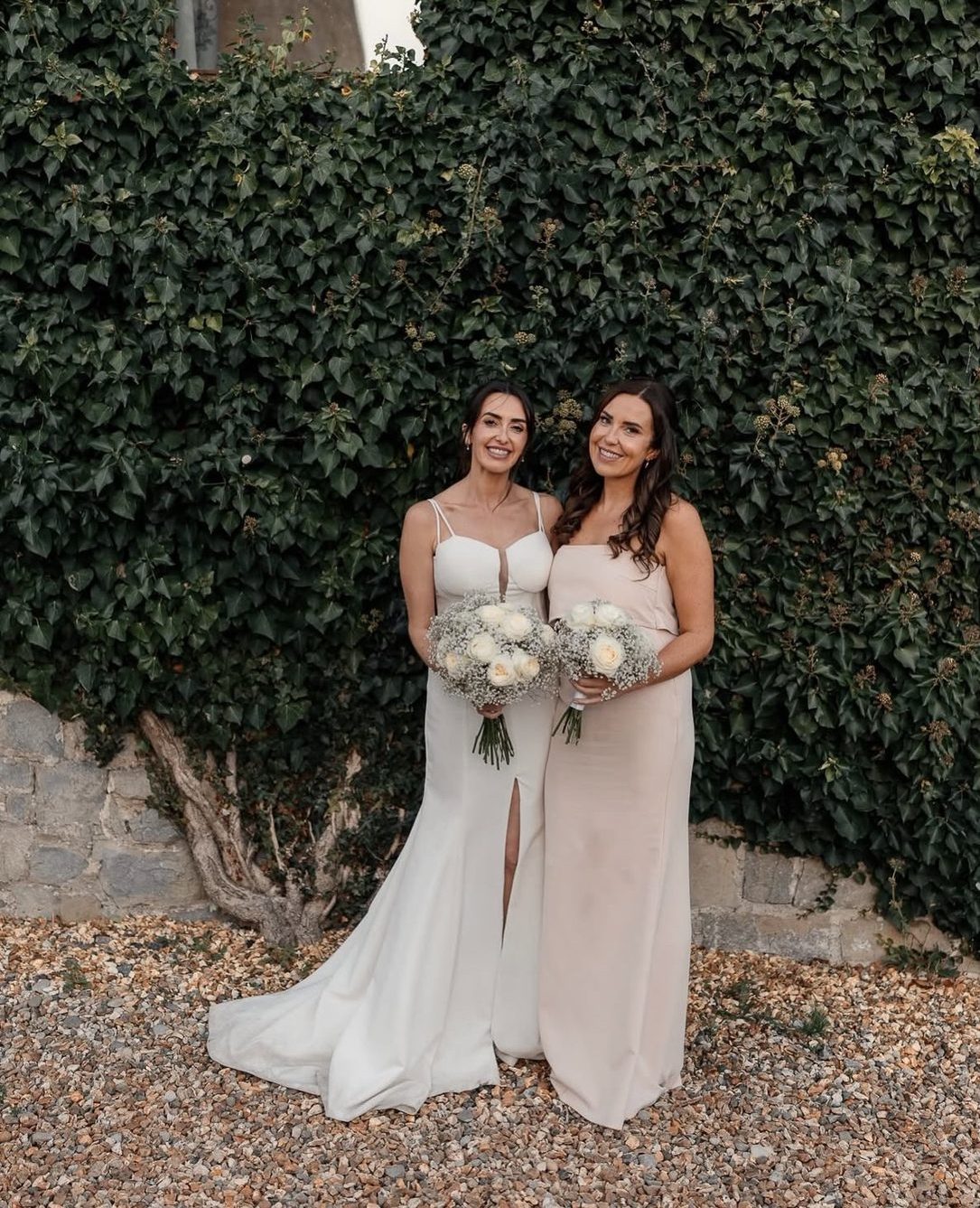 Bride and bridesmaid holding white bouquets against an ivy wall